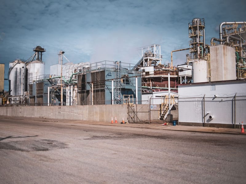 An industrial plant exterior with silos, pipework and access gantries behind a fence