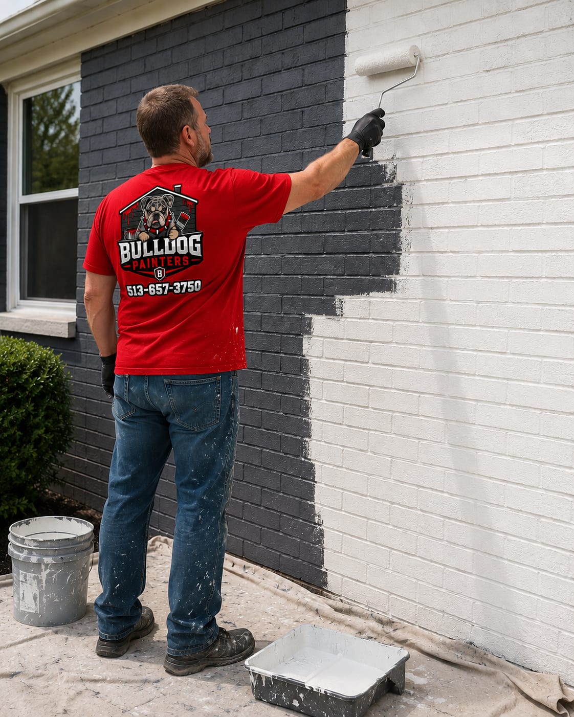 A Bulldog Painters painter rolling white masonry paint over dark brick, half the wall still unpainted