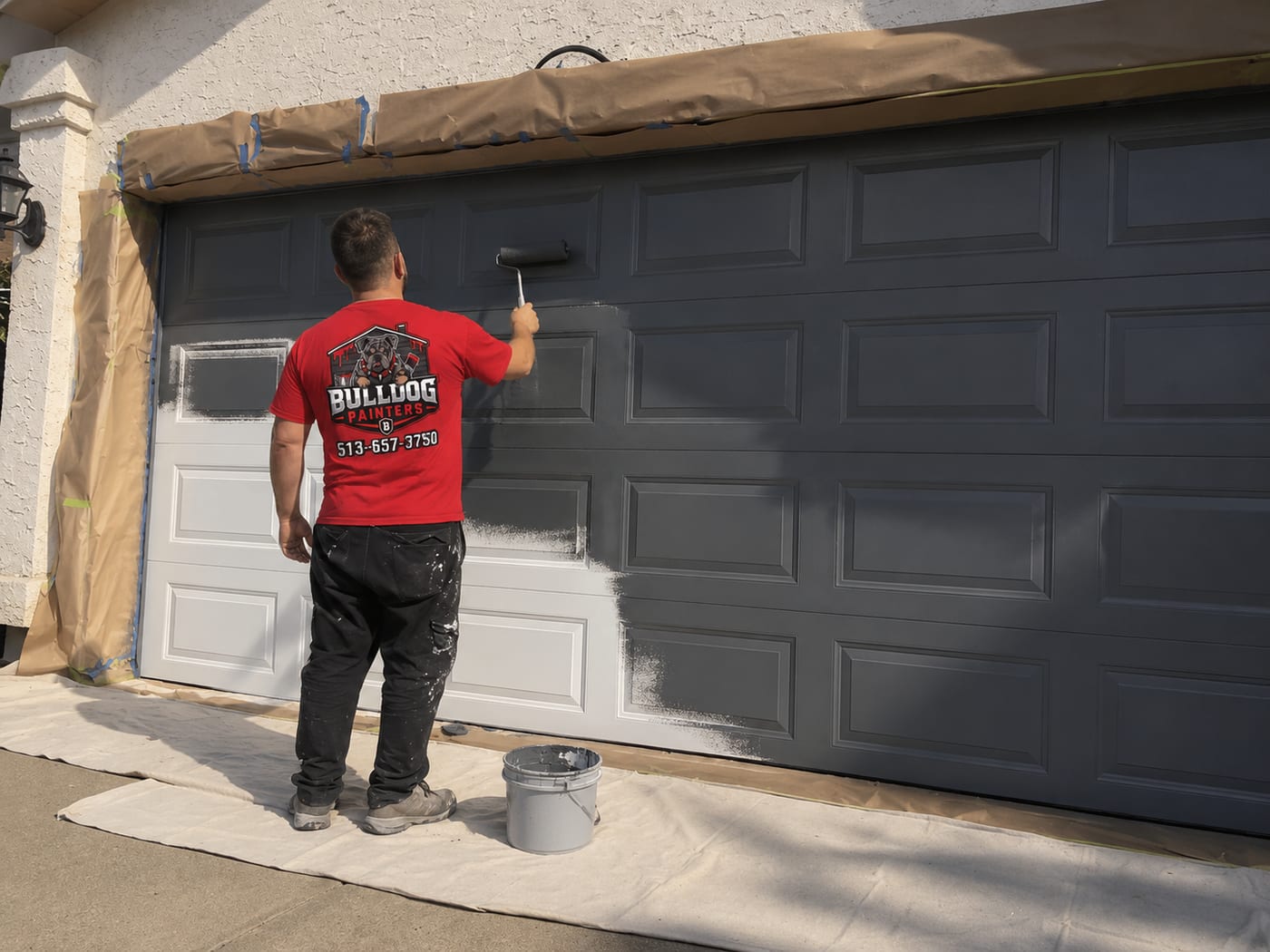 A Bulldog Painters painter rolling a garage door, drop cloths down across the driveway