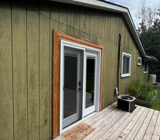 A house exterior before painting — olive-stained board siding with bare wood trim around the patio door