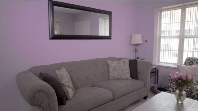 A living room painted pale lilac, with a grey sofa below a long mirror and a window to the right
