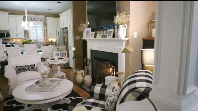 A living room with a white painted fireplace and mantel, an open white kitchen behind it, and a black and white checked armchair in the foreground