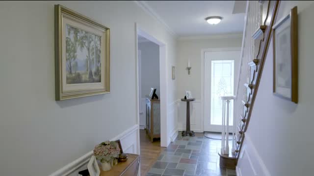 An entry hall painted pale grey-green with white wainscot and crown molding, a framed landscape on the left, a slate floor by the front door and a staircase with white balusters on the right