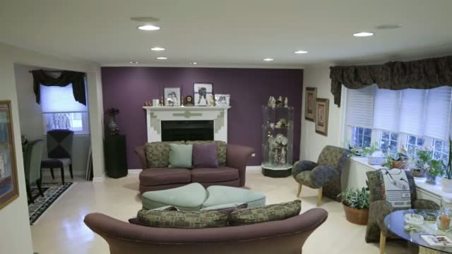 A living room with a plum accent wall behind a white painted fireplace, pale walls and white trim to either side, and a bay window on the right